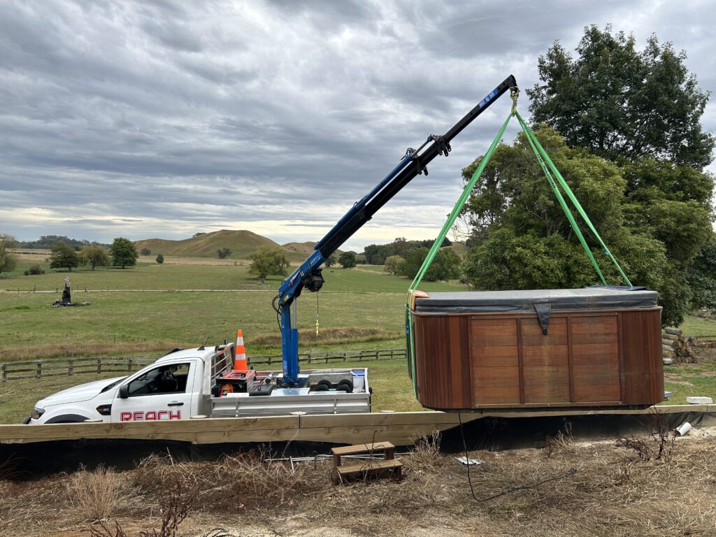 A mini Hiab crane lifting a wooden spa pool over rural paddocks for precise placement onto a residential deck.