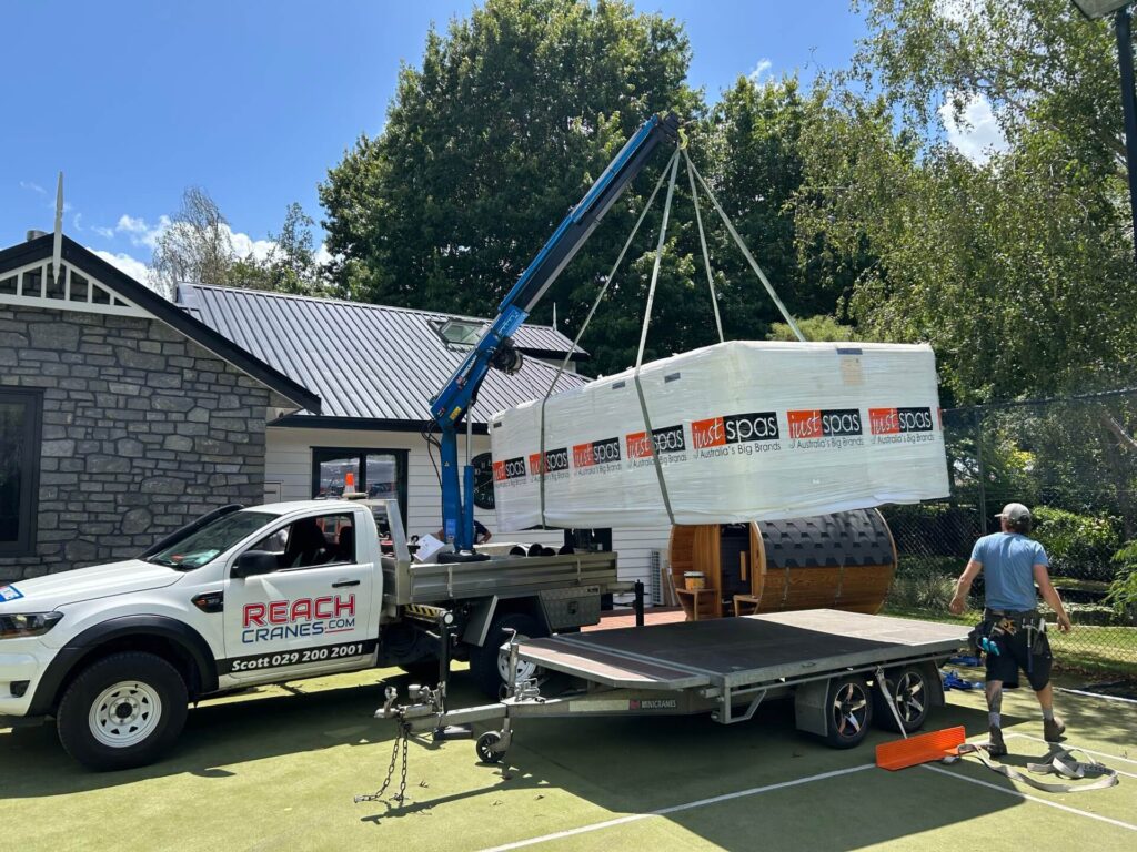 A wrapped swim spa being precisely lifted off a tennis court by Reach Cranes’ Hiab hire, with expert movers guiding the lift step by step.
