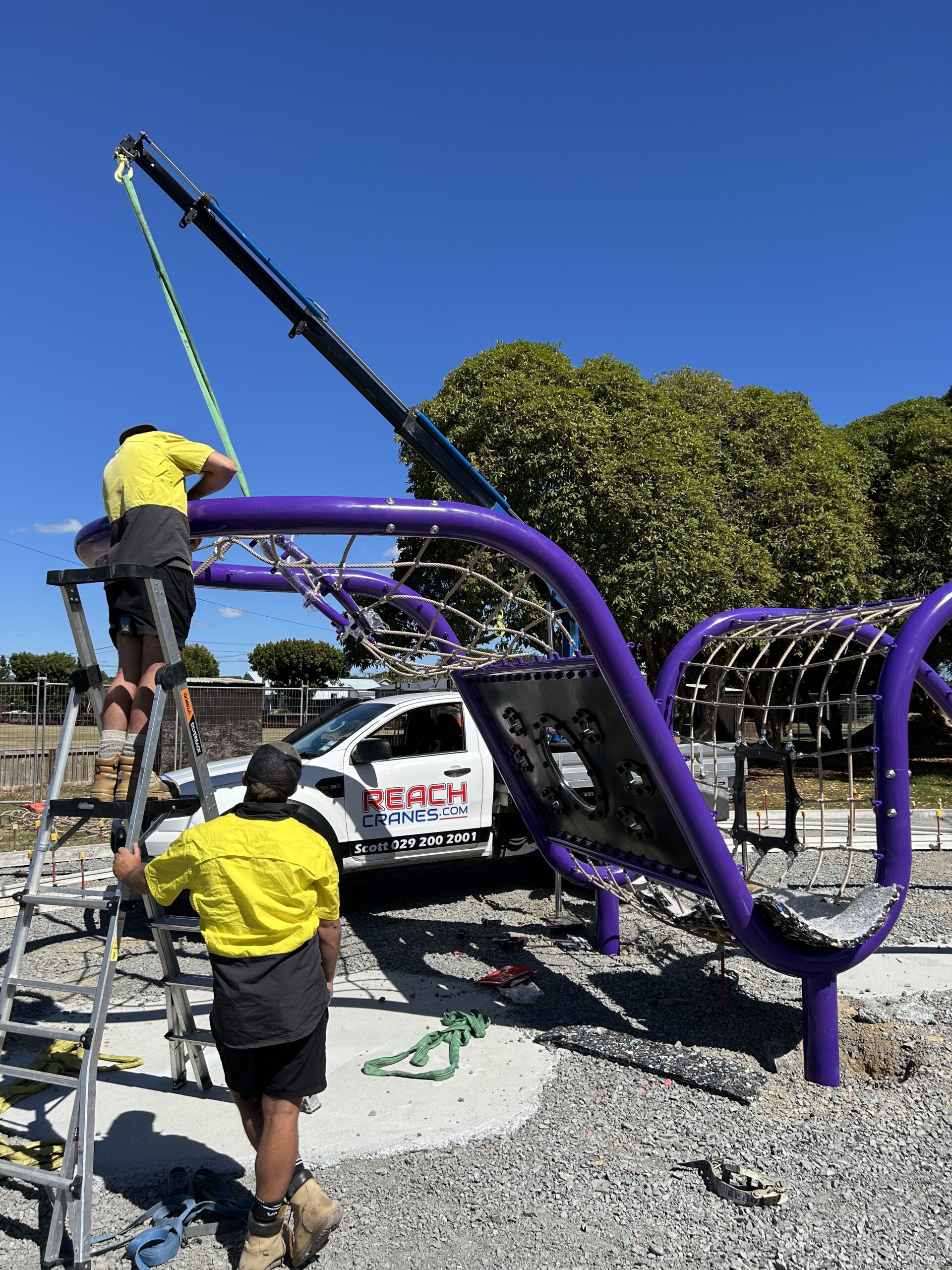 A Reach Cranes mini crane lifting a section of purple climbing playground equipment into place, supported by on-site installers.