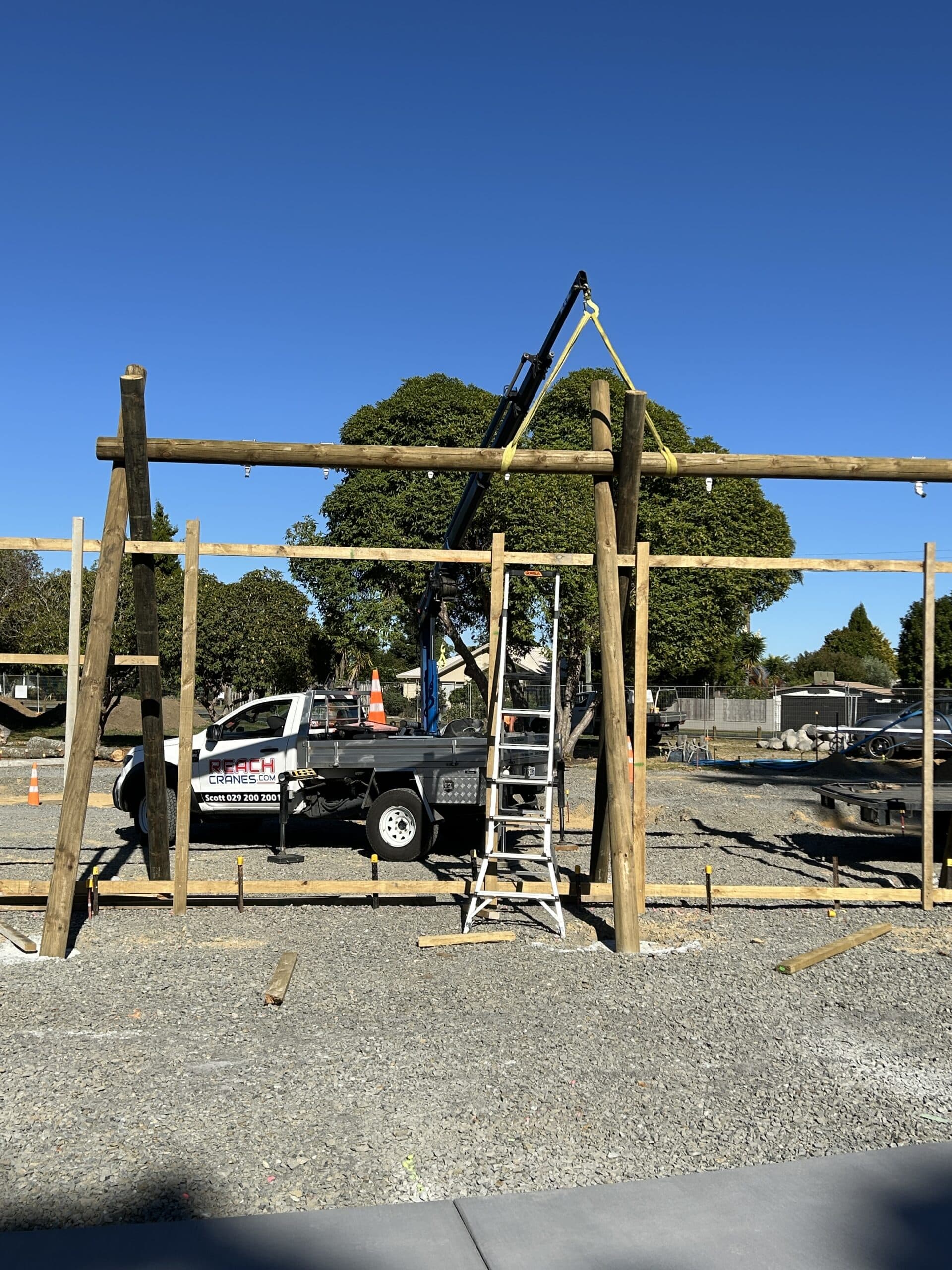 A Reach Cranes mini crane hoisting large timber swing frame poles during a playground installation project.