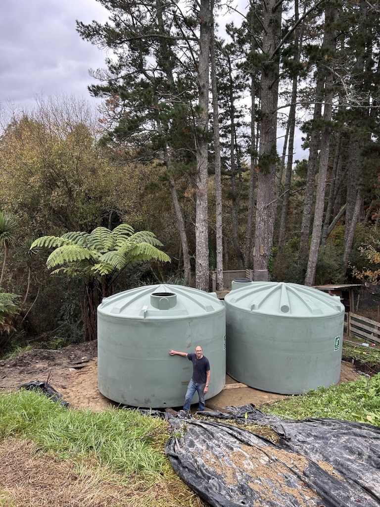 A satisfied client standing beside two large green water tanks successfully delivered and installed on a steep rural site near Raglan Beach, surrounded by native bush and pine trees.