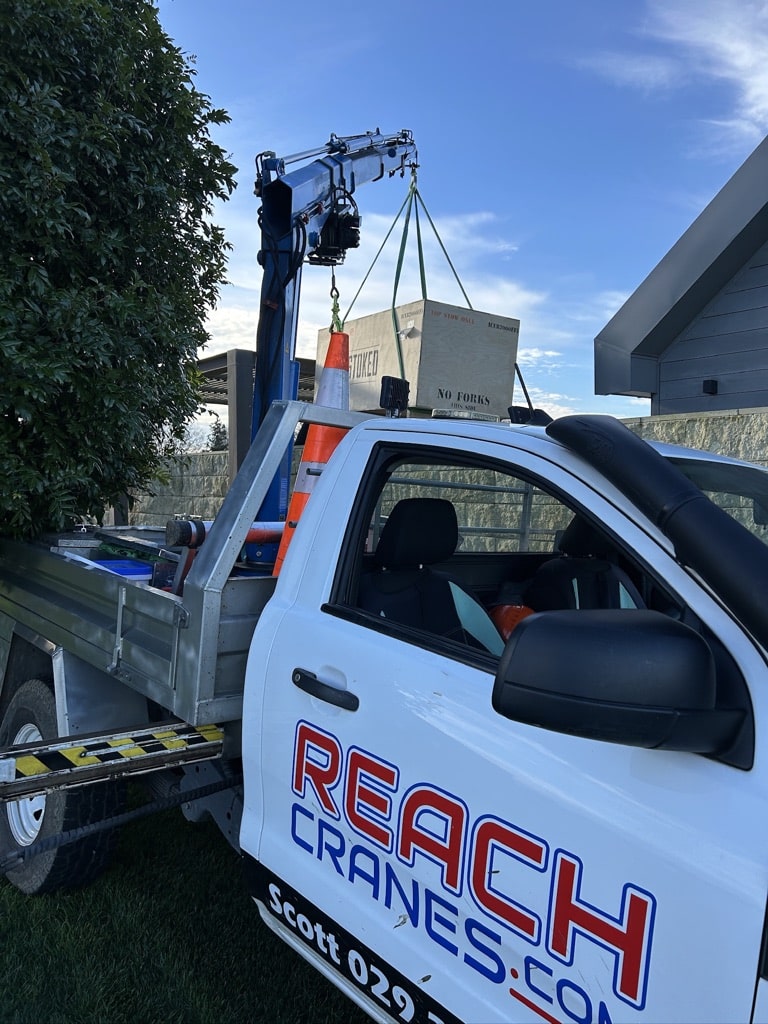 A Reach Cranes mini Hiab crane lifts a Stoked Stainless spa pool crate over a fence for residential delivery.